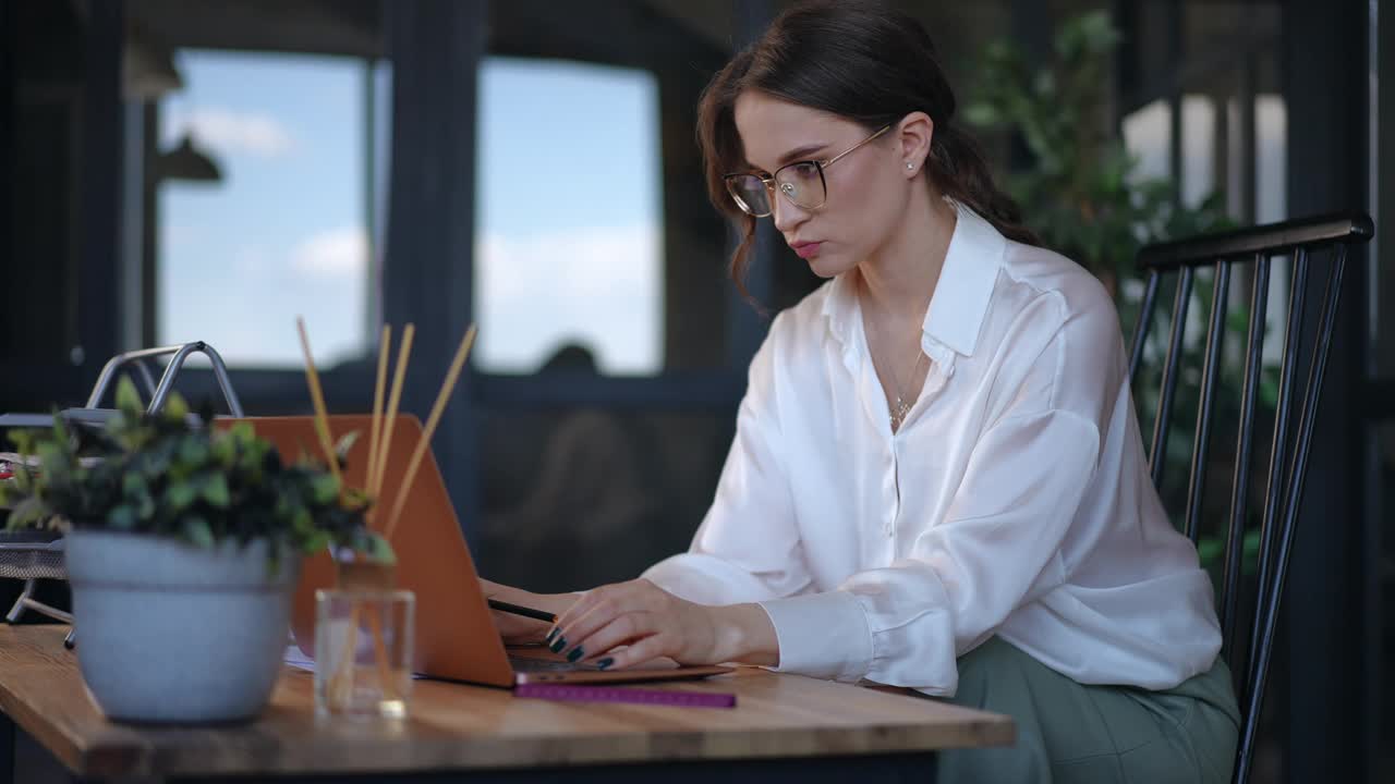 一位漂亮的女商人戴着眼镜，穿着白衬衫，坐在现代办公室木桌前的椅子上，用笔记本电脑工作。一位女士在笔记本电脑键盘上打字，若有所思地看着。视频素材