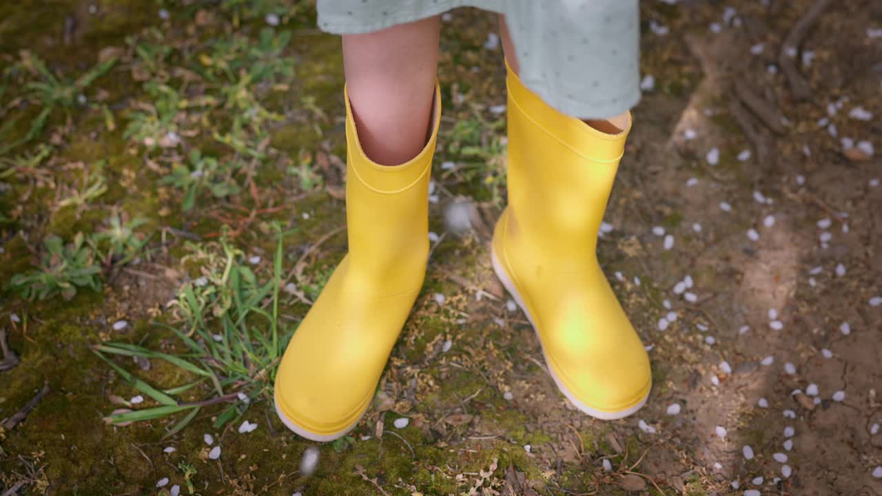 一个美丽女孩的黄色农妇靴和花园中绿草和农田上盛开的春天树木的花瓣视频下载