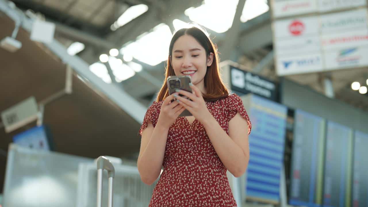 微笑的亚洲女子在机场用智能手机发短信，在旅行中进行友好的聊天视频下载