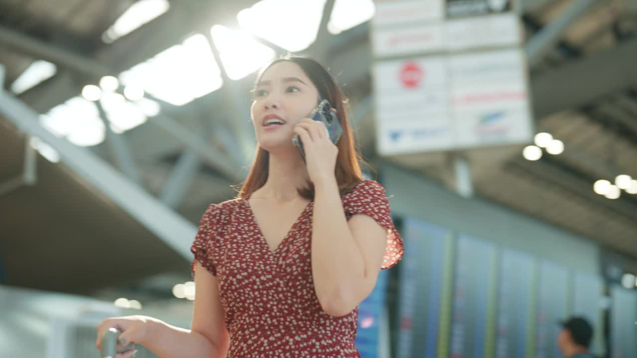 快乐的年轻亚洲女子在机场航站楼一边拉箱子一边聊手机视频下载
