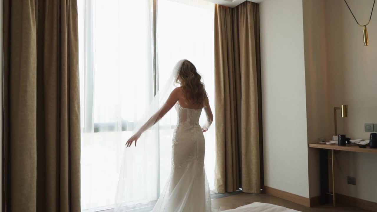 后台婚礼拍摄。身着婚纱、头戴面纱的新娘站在窗边。视频下载