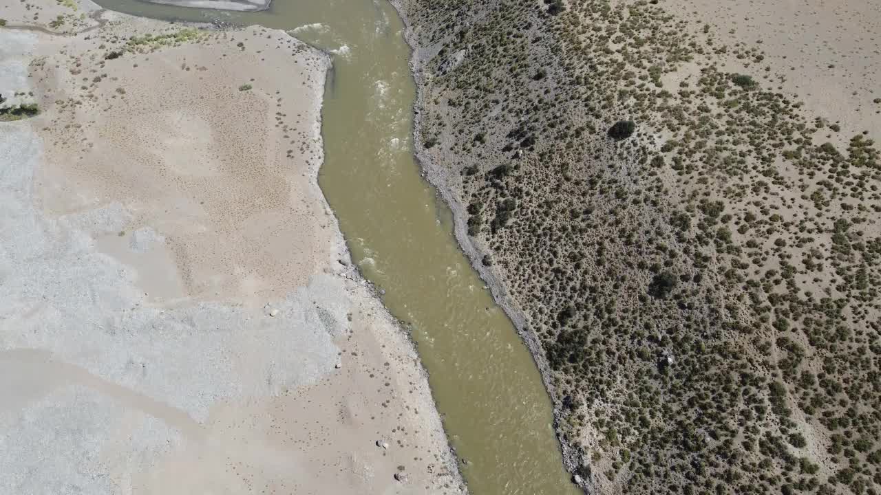 镜头沿着一条小河向上平移，棕色的水穿过安第斯山脉脚下的干旱景观，远处有山脉。视频下载