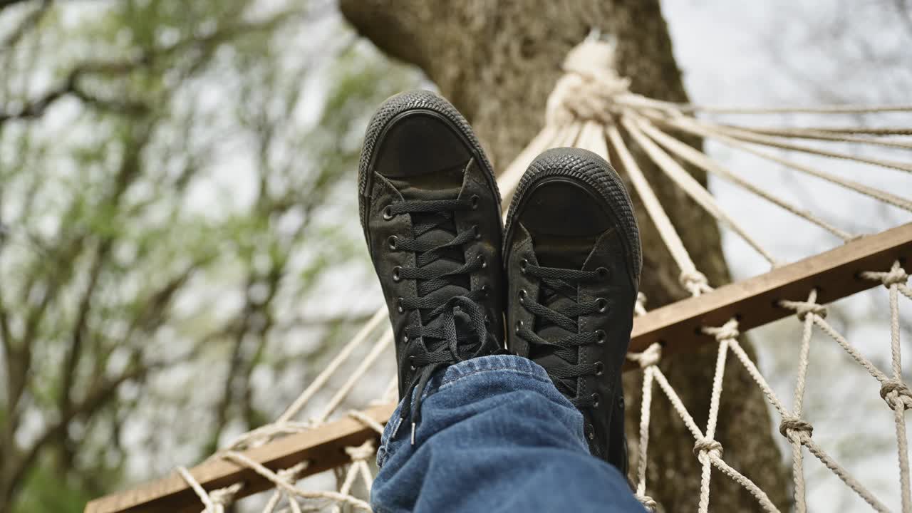 一名男子穿着牛仔裤和脏兮兮的黑色运动鞋，躺在后院树上的吊床上休息视频下载