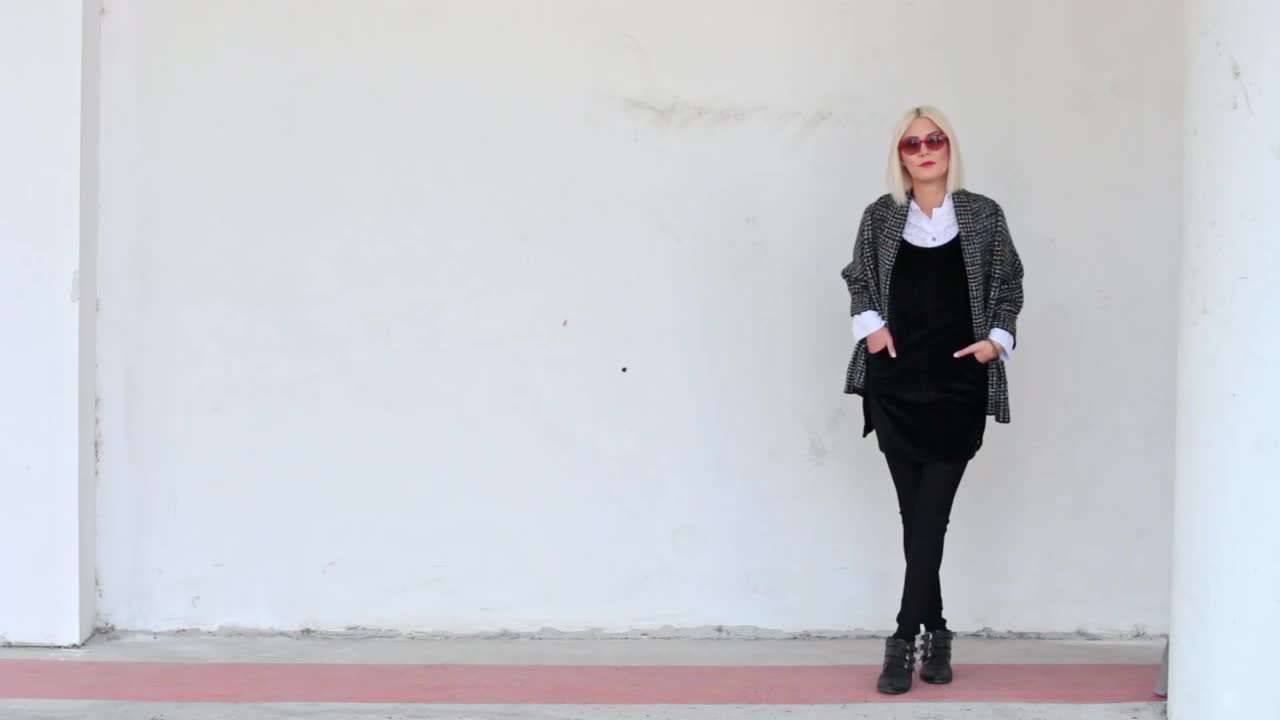 金发模特穿着黑白相间的衣服，戴着红色太阳镜站在白色的墙前视频下载