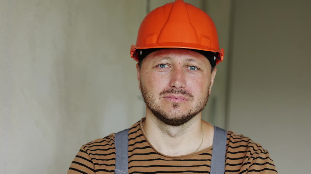装修公寓。大胡子的自信的专业维修承包商的肖像，戴着安全的橙色安全帽和工作服，看着镜头。空的混凝土墙，修理房子，新建大楼视频下载