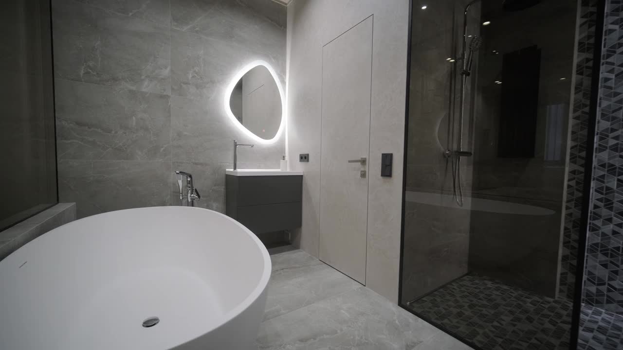 从卧室看时尚而现代的浴室，展示了带背光的椭圆形镜子，一个独立的浴缸和一个带几何瓷砖的步入式淋浴间。视频下载