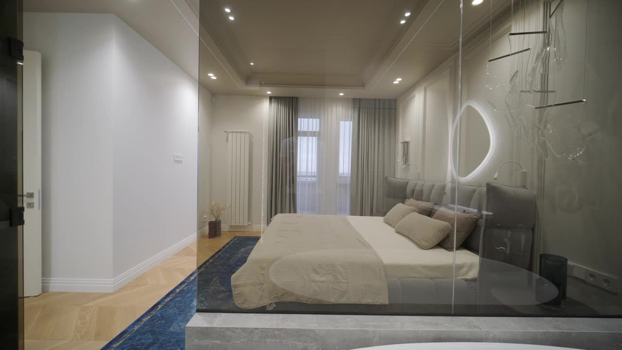 一间豪华的卧室，配有时尚的簇绒床头板和醒目的蓝色地毯，无缝过渡到一间带有现代灯具和独特椭圆形镜子的时尚浴室，捕捉了融合舒适与优雅的设计。视频下载