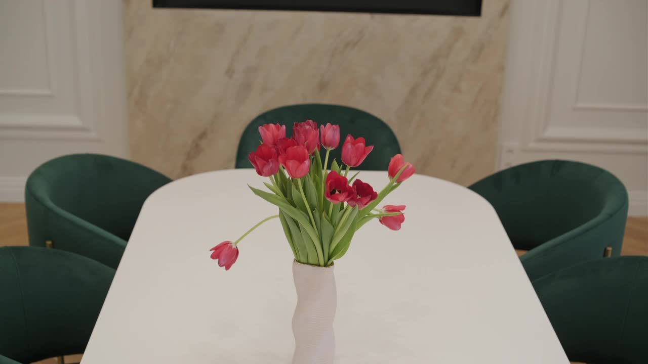 光滑的白色桌子上放着一个白色花瓶，里面放着一束鲜红的郁金香，背景是绿色的天鹅绒椅子视频下载
