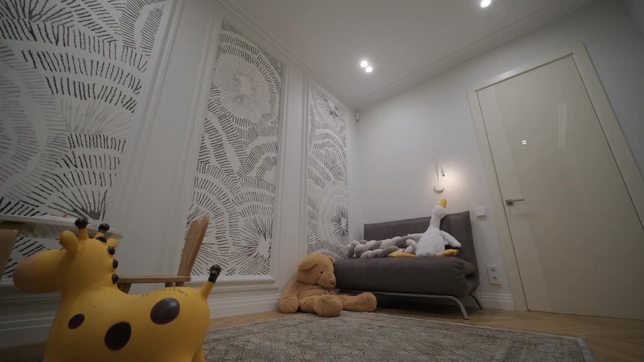 一个舒适的儿童房角落里，有一个好玩的长颈鹿玩具和一个可爱的泰迪熊，旁边是一个小灰色沙发，衬着异想天开的图案壁纸，创造了一个迷人和养育的空间视频下载
