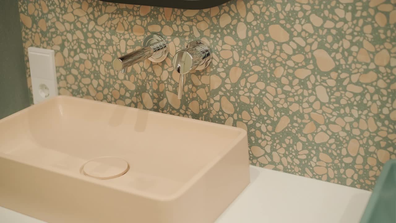 这是一个别致的浴室角落的细节照片，一个桃红色的面盆与水磨石瓷砖的墙壁和抛光的镀铬水龙头相衬，传达出一种现代和艺术气息视频下载