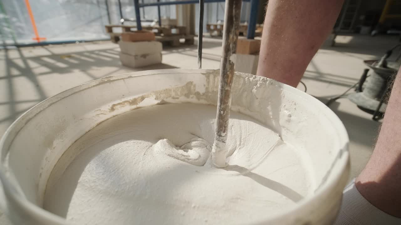 一个建筑工人的腿在一个水桶旁边，他用混合工具混合一批白色石膏，在地面上投下阴影视频下载