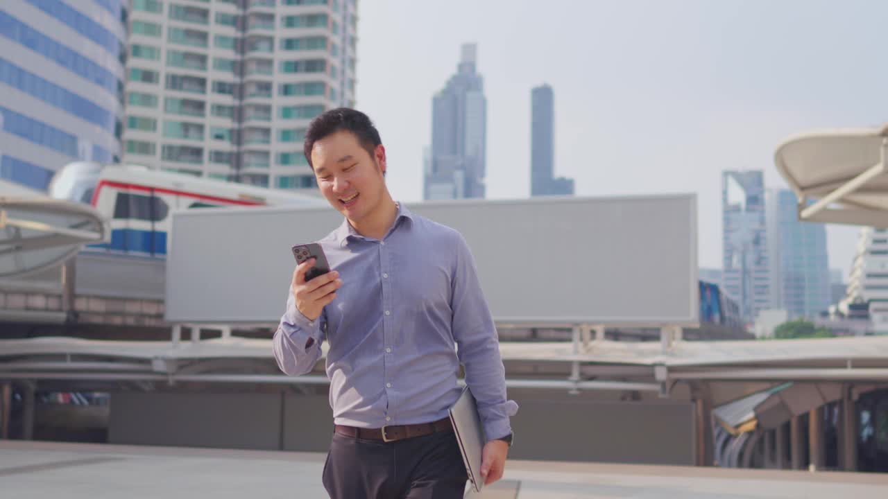 亚洲商人在城市户外使用数字智能手机进行通信。有魅力的男员工心情愉快，在现代化的镇中心边走边打电话。视频下载