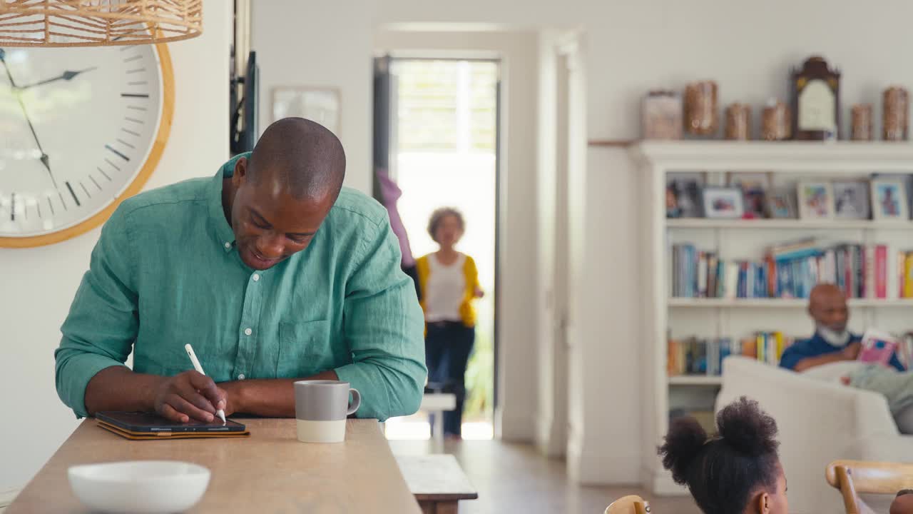 一名男子在室内用触控笔操作数字平板电脑，背景是多代同堂的家庭成员视频下载