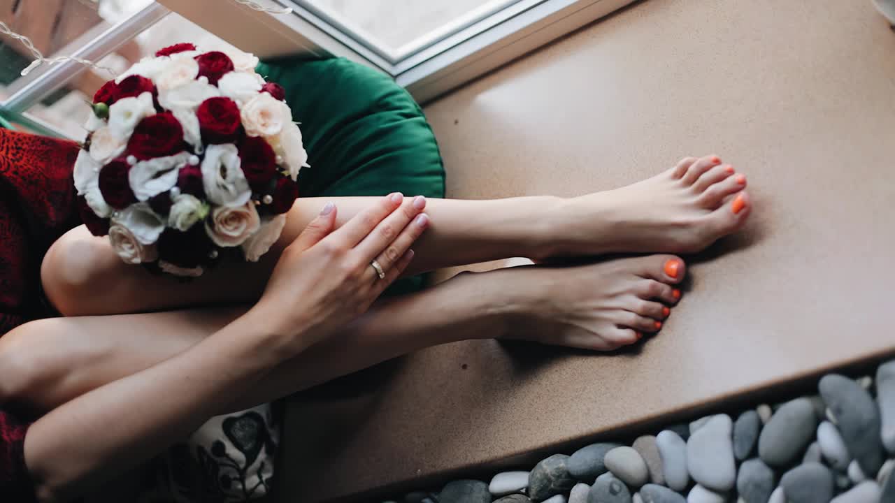 一个女孩坐在窗台上抚摸着她的腿。她手里拿着一束鲜花。从上方拍摄视频下载