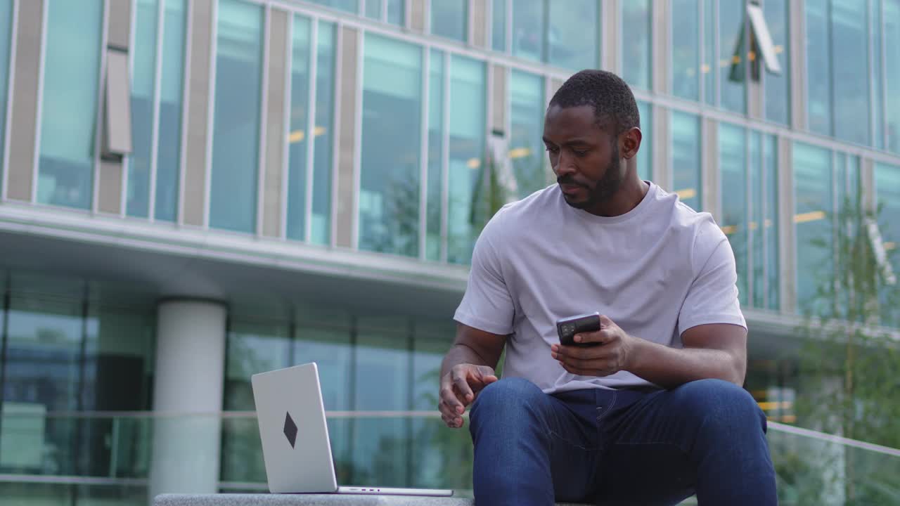 非裔美国自由职业者在城市街道上用笔记本电脑打电话。一个商人正在开虚拟会议。专业程序员使用手机和笔记本电脑在户外工作。远程工作。视频下载