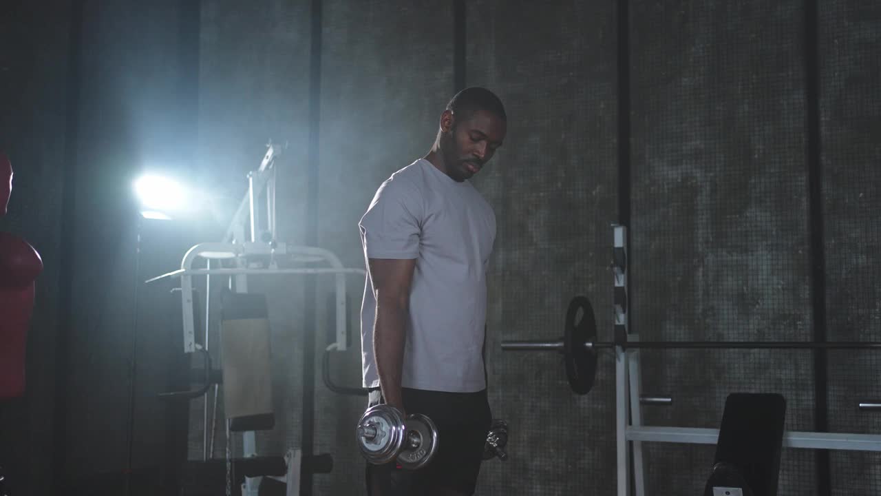 在健身房健身。非裔美国男子健美运动员在健身房拿哑铃。在健身俱乐部用哑铃锻炼二头肌肌肉。男子在做体育锻炼。健康的生活方式视频下载
