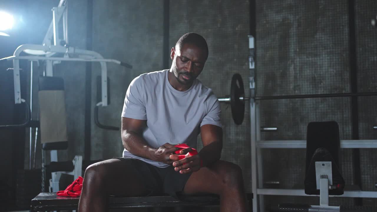健身馆里的健身训练。非裔美国男子拳击手在健身馆里用红色的拳击绷带包裹着运动保护绷带。强壮的男子准备进行拳击对练训练。视频下载