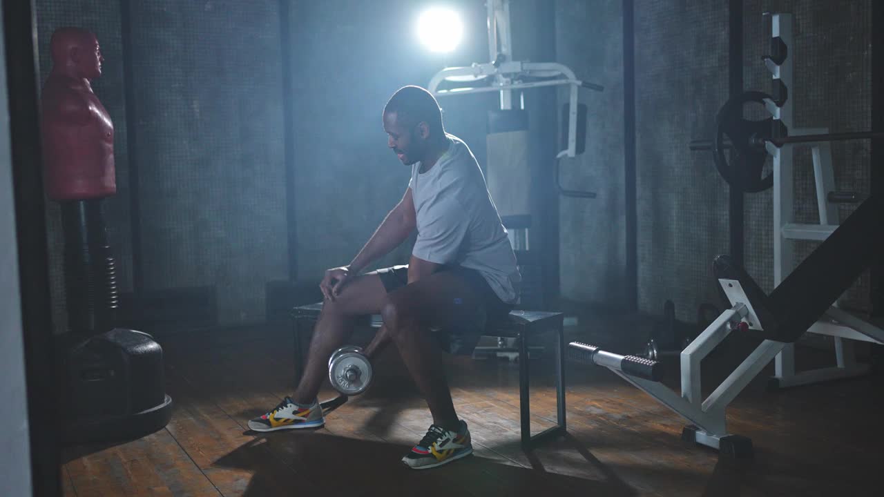 在健身房健身。非裔美国男子健美运动员在健身房拿哑铃。在健身俱乐部用哑铃锻炼二头肌肌肉。男子在做体育锻炼。健康的生活方式视频下载