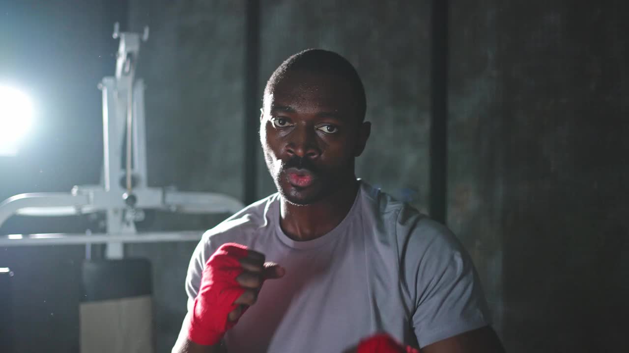 在健身房锻炼。非洲男子拳手准备打拳击，对着镜头裹上保护绷带。强壮的男子训练出拳，看起来很集中，正准备拳击对练。视频下载