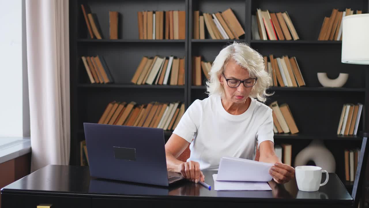 自信时尚的中年女性在工作场所使用笔记本电脑。年长成熟的女商人坐在办公桌旁，手里拿着笔记本电脑和纸质文件。老板、领导、老师、专业工作者。视频下载
