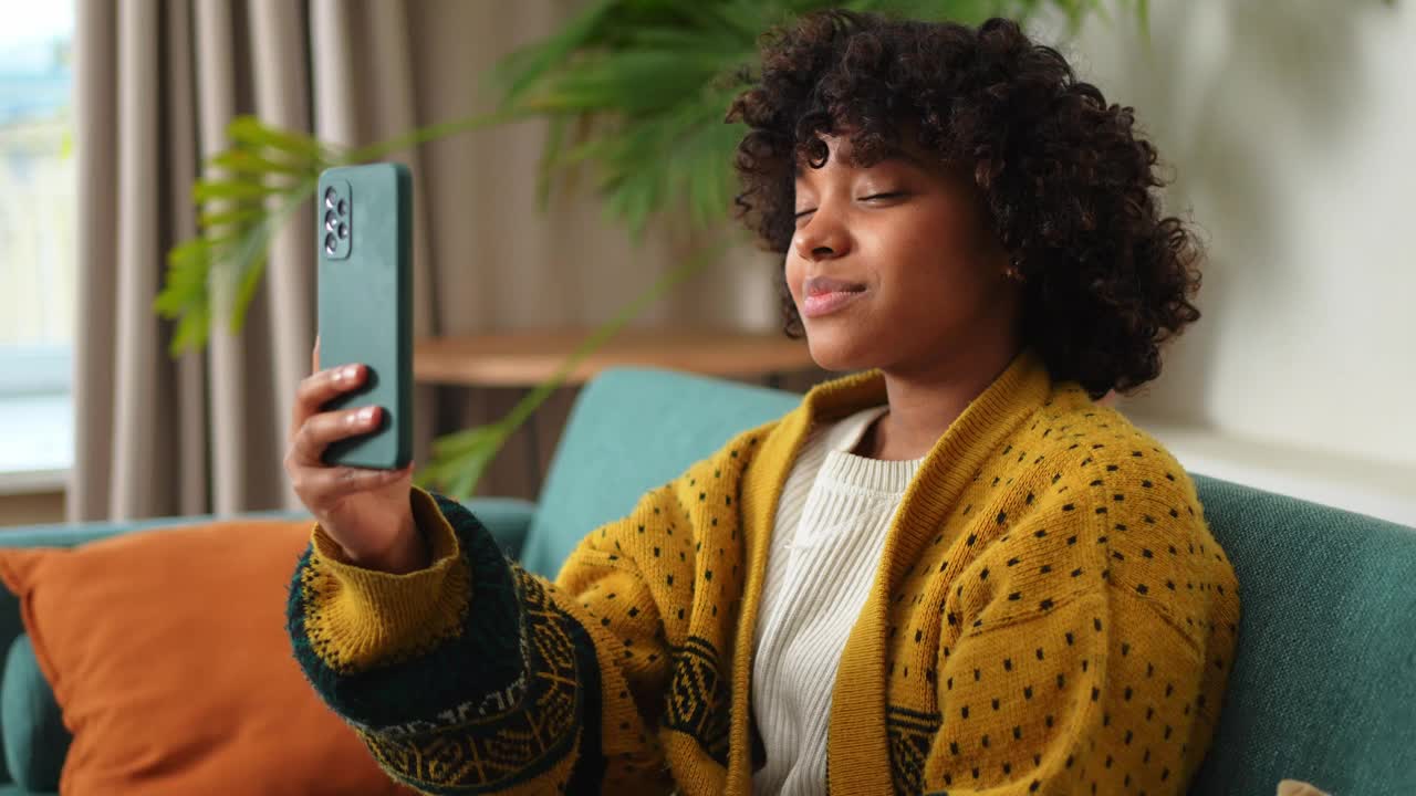 非裔美国女孩拿着智能手机视频聊天。女博主在社交网络上与最好的朋友聊天。年轻女子在家里进行虚拟会议在线聊天视频通话。视频下载