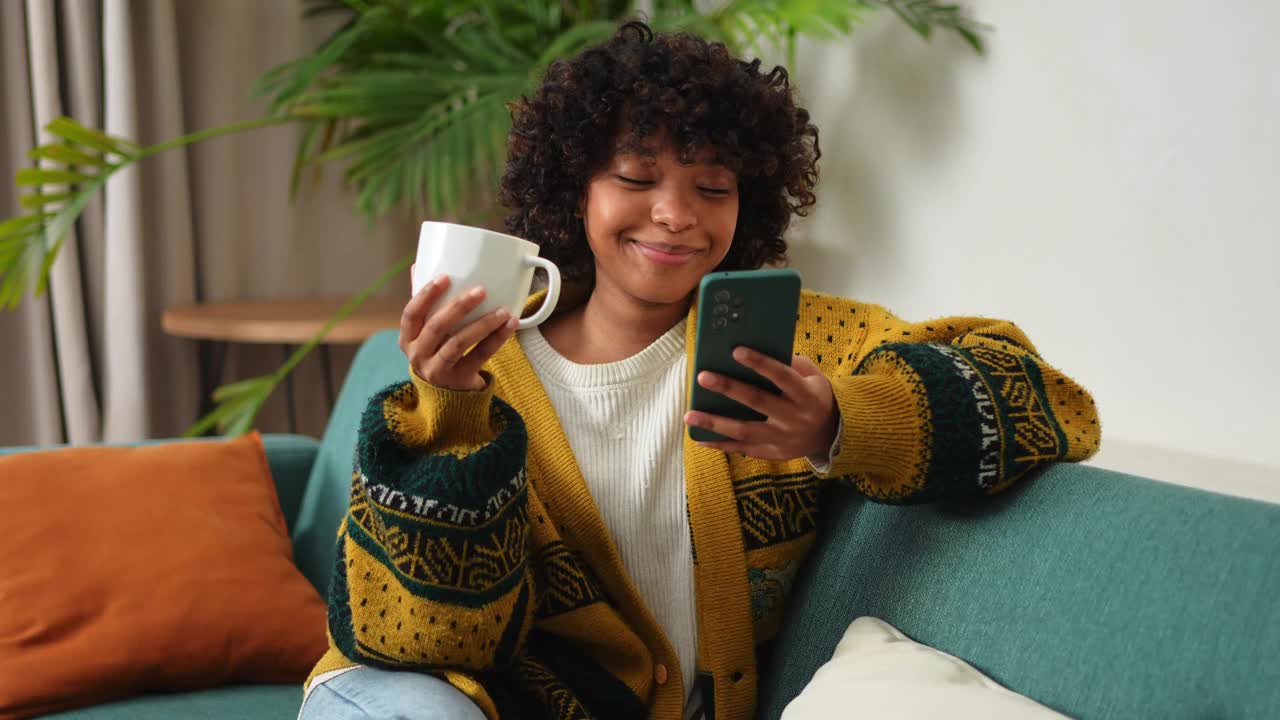 早上好，非洲女孩喝咖啡，拿着智能手机坐在家里的沙发上，女人用手机上网，使用社交媒体应用程序。上网购物上网新闻手机成瘾视频下载