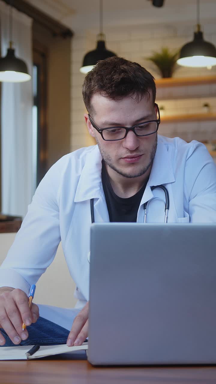 垂直视频。专业的年轻男医生戴着眼镜，穿着白色制服，戴着听诊器，通过笔记本电脑的网络摄像头与患者进行在线会议视频通话。远程医疗咨询视频下载