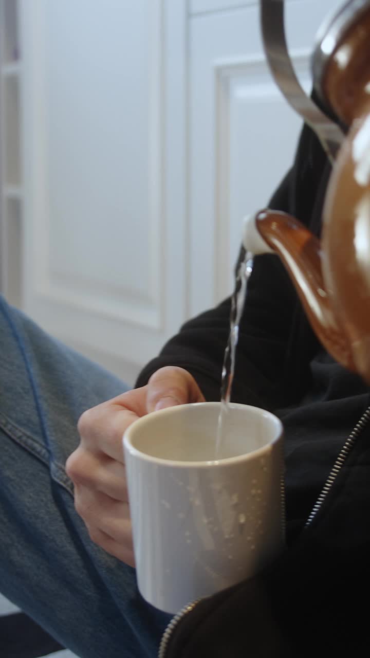 垂直视频。一个年轻人坐在厨房的地板上，从茶壶里往杯子里倒满了水和饮料。视频下载