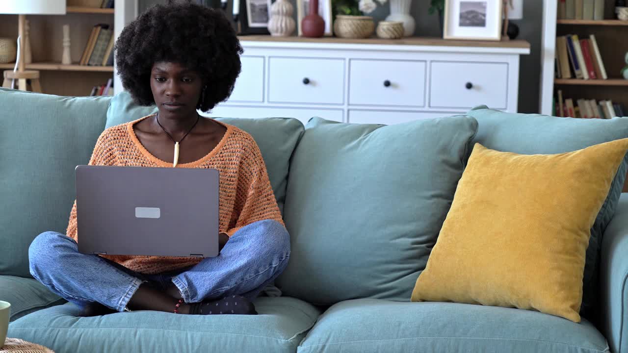 一位年轻的非裔美国女性在沙发上放松地使用笔记本电脑视频下载