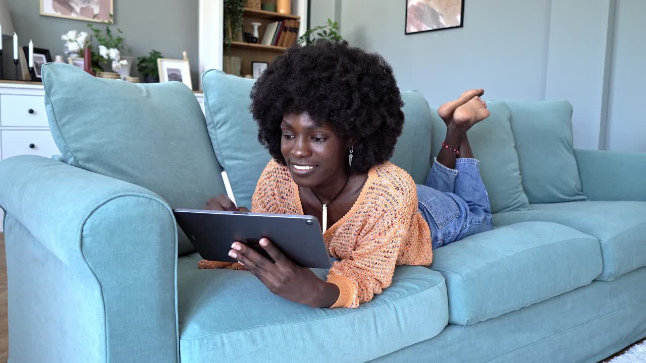 开朗的年轻女子在沙发上放松，同时在家使用数码平板电脑视频素材