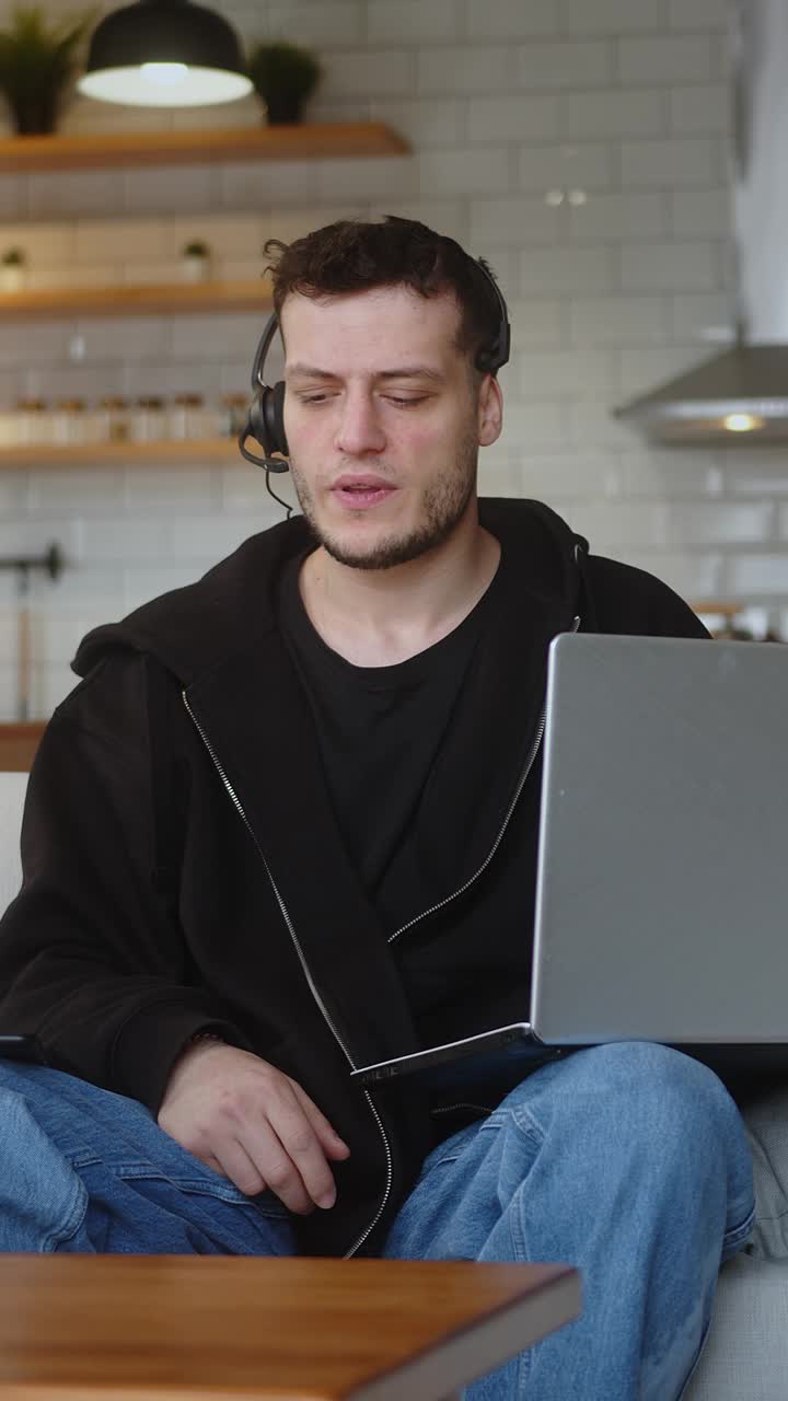 垂直视频。呼叫中心的男代表戴着无线耳机，用笔记本电脑帮助客户投诉。公司操作员坐在沙发上远程办公，为客户提供支持。视频下载