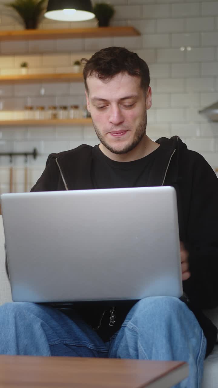 垂直视频。微笑的年轻人坐在客厅的沙发上用笔记本电脑工作。上网，用马克杯喝咖啡，在家里玩。视频下载