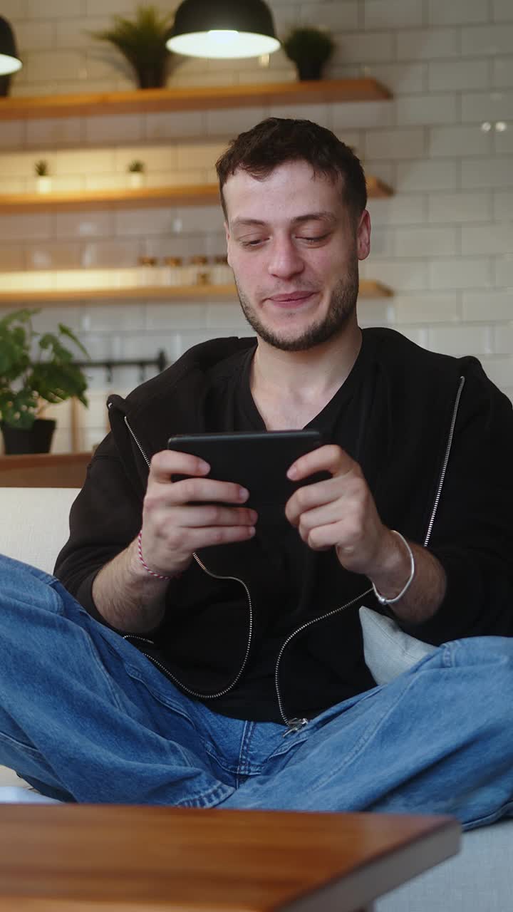 垂直视频。一个专注的年轻人坐在家里的沙发上玩着智能手机上的电子游戏。兴奋集中的人全神贯注于有趣的游戏，独自享受乐趣视频下载