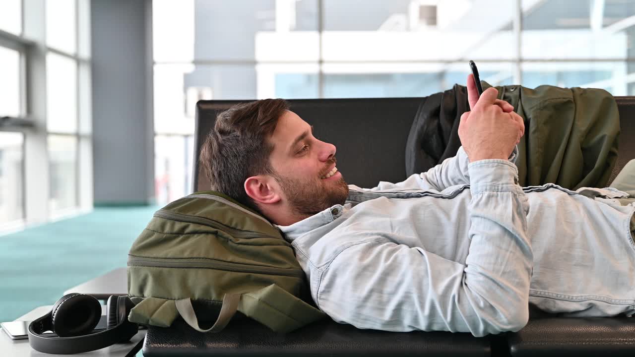千禧一代商务旅客在机场躺着等待时使用智能手机和互联网数据视频下载