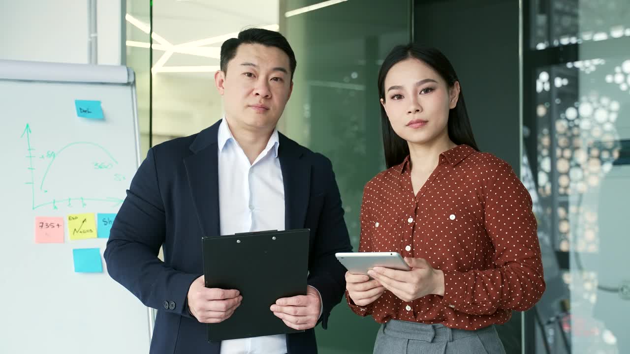 幸福的亚洲男经理和女员工站在办公室里，拿着笔记本电脑和数码平板电脑看着相机。视频下载