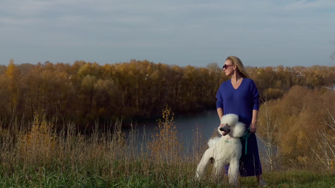 秋天，一名女子牵着狗绳在河边遛狮子狗视频下载
