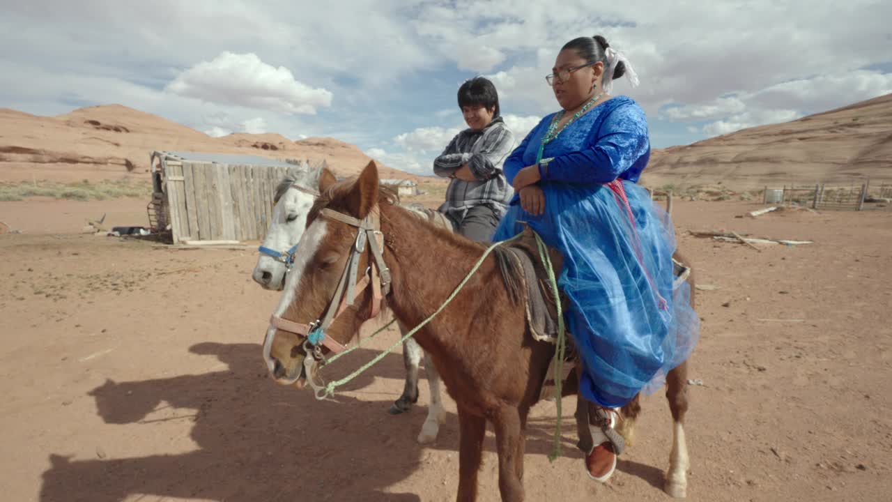 20多岁的年轻纳瓦霍夫妇在纪念碑谷部落公园的家庭牧场上骑马，背景是美丽的岩层视频下载