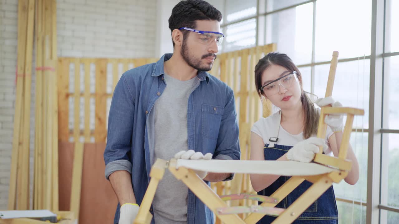 快乐的年轻男木匠和女木匠戴着棉手套和护目镜，用黄胶带测量支撑椅墙的长度，一起在木匠店里组装椅子，制作新设计的木椅。视频下载