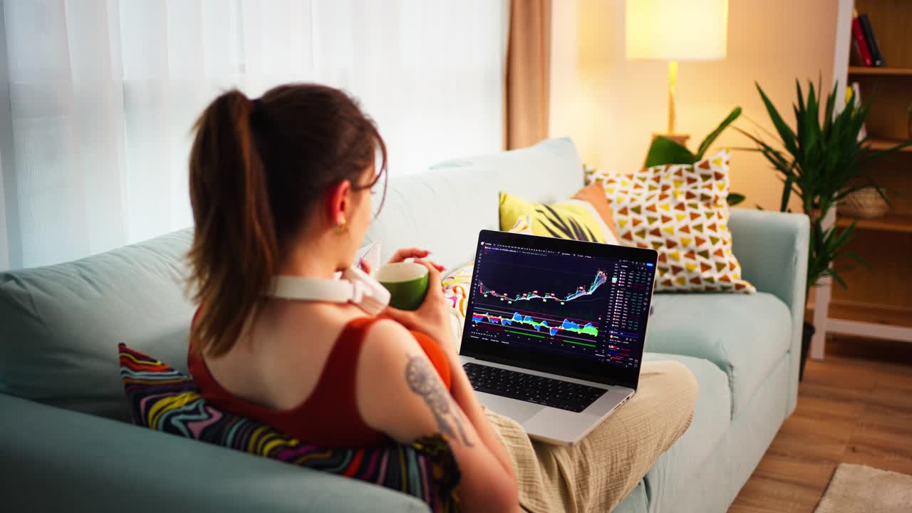年轻的女商人坐在沙发上，伸开双脚，用笔记本电脑显示器和手机在数字交易所查看比特币或股票的价格走势图视频下载