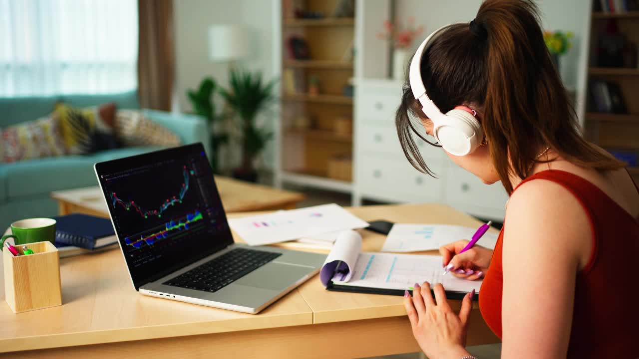 戴着耳机的年轻女商人在家里桌上的笔记本电脑上查看数字交易所的比特币或股票价格图表视频下载