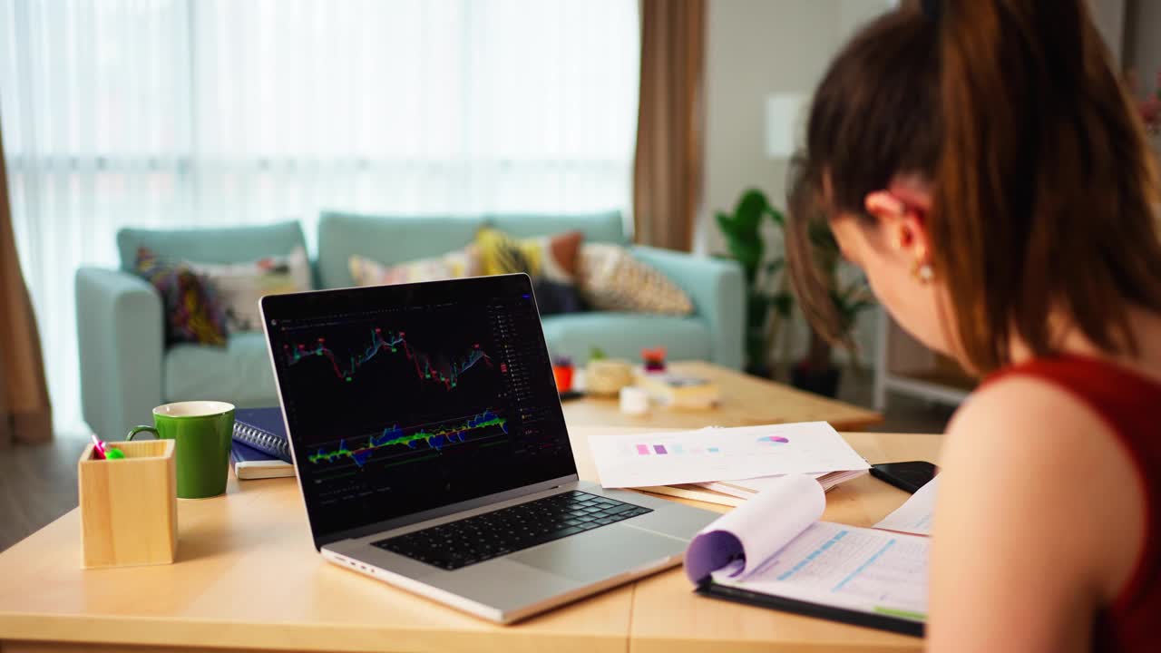年轻的女商人在家里的桌子上用笔记本电脑查看数字交易所的比特币或股票市场价格图表，视频下载