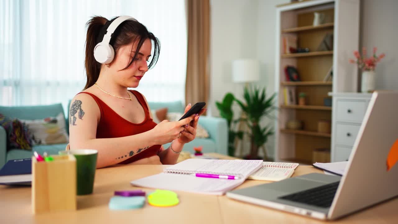 年轻的女商人，大学生，在家办公室用笔记本电脑工作时，一边用手机视频下载