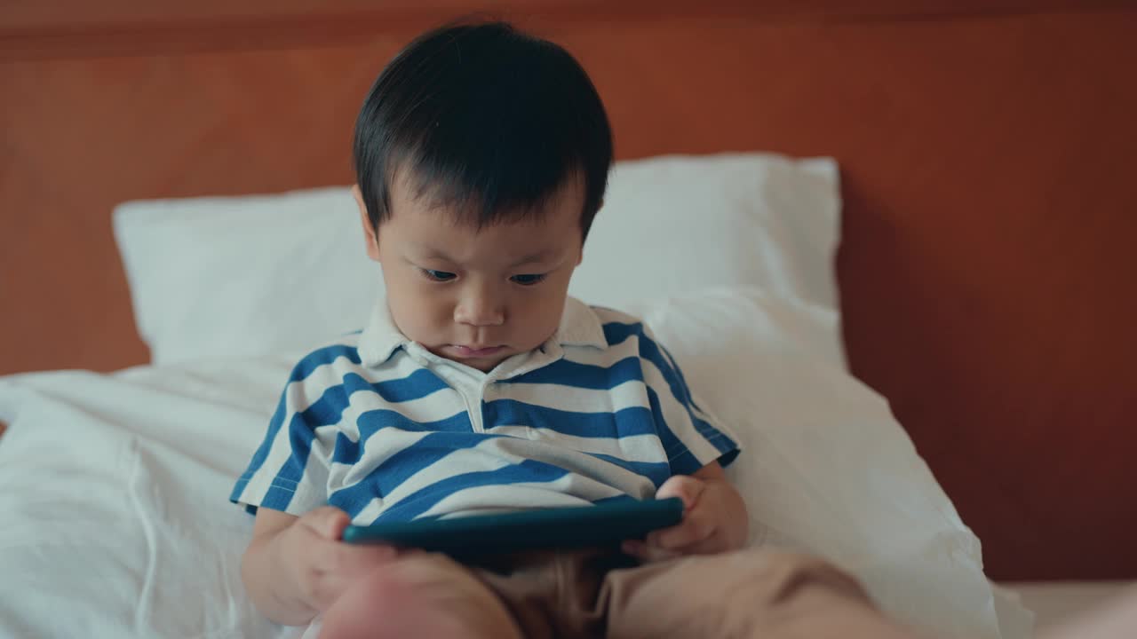 亚洲可爱的小男孩坐在床上，在家用智能手机。视频下载