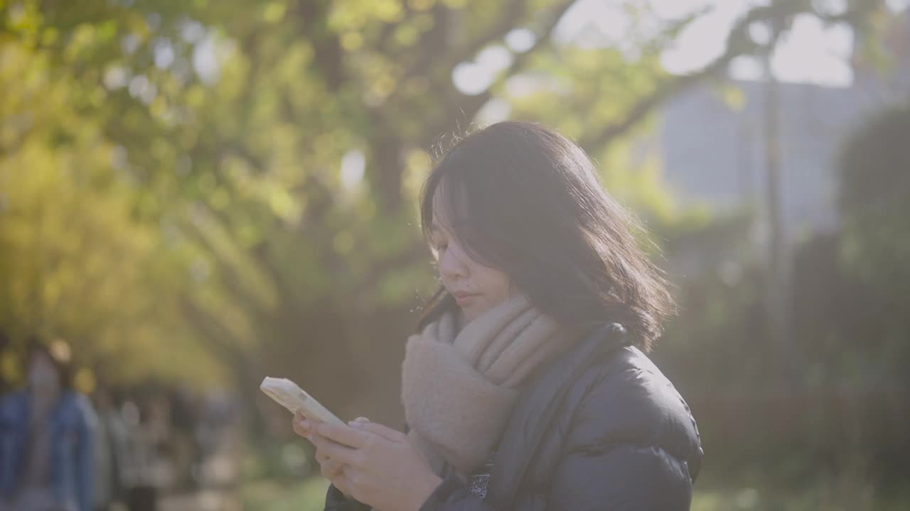 一个寒冷的秋天，年轻的亚洲女子在公园里放松。她通过智能手机拍摄了一张令人惊叹的照片或视频。视频下载