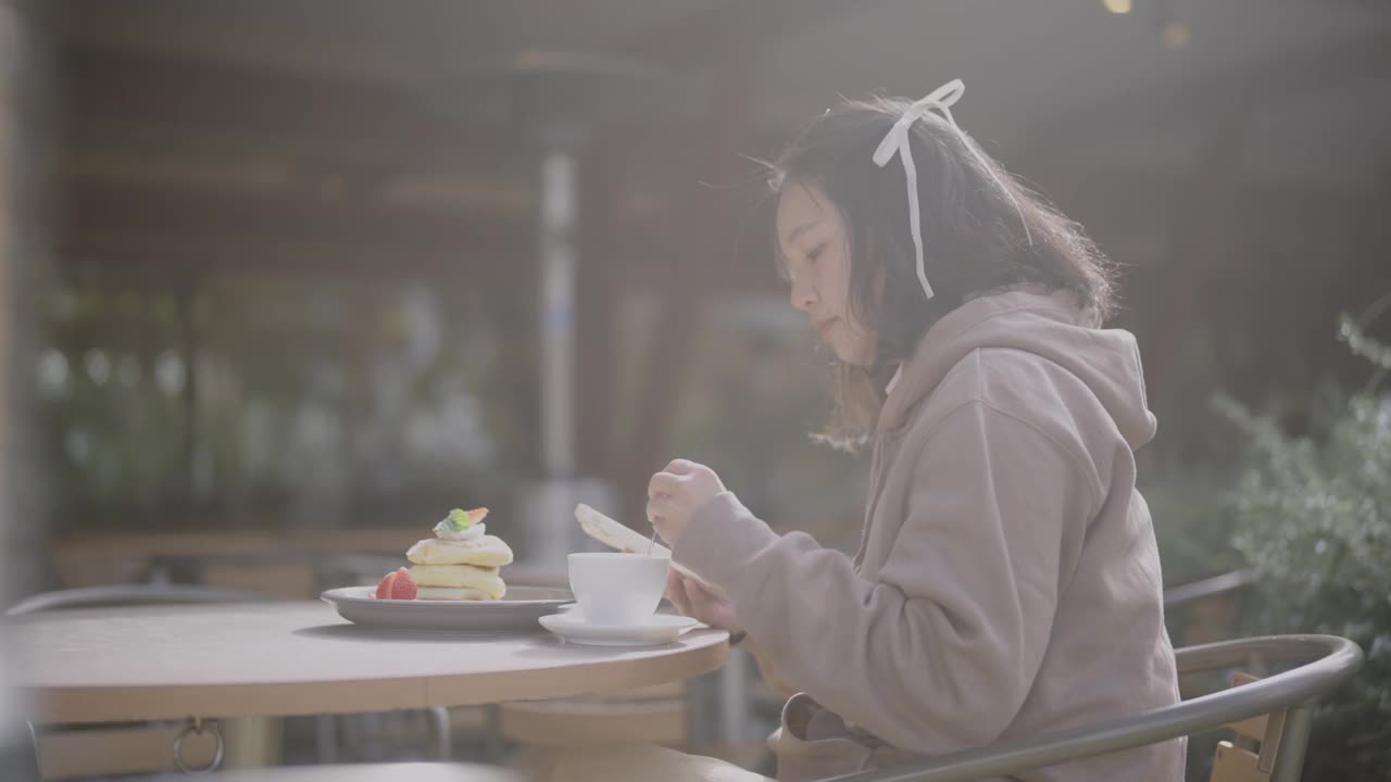 一个寒冷的秋天，年轻的亚洲女子在咖啡馆里放松。她在晴天吃煎饼。视频下载