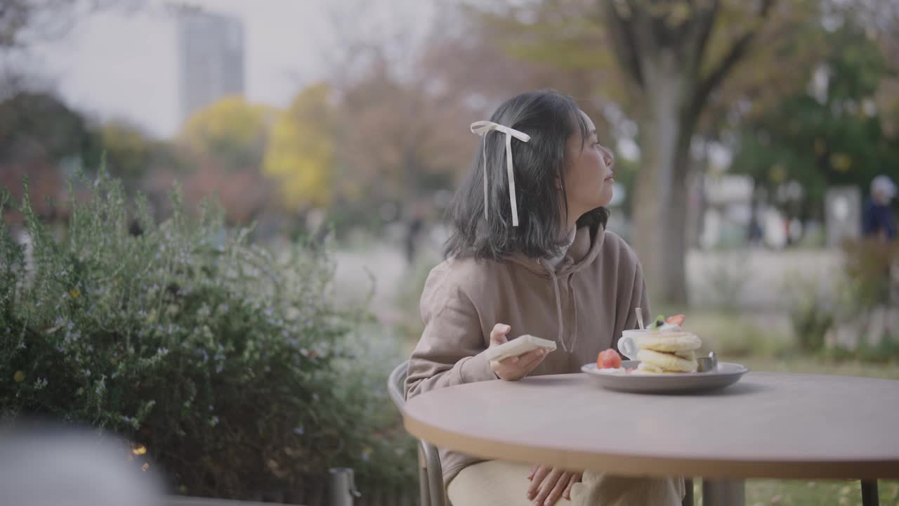 一个寒冷的秋天，年轻的亚洲女子在咖啡馆里放松。她在晴天吃煎饼。视频下载
