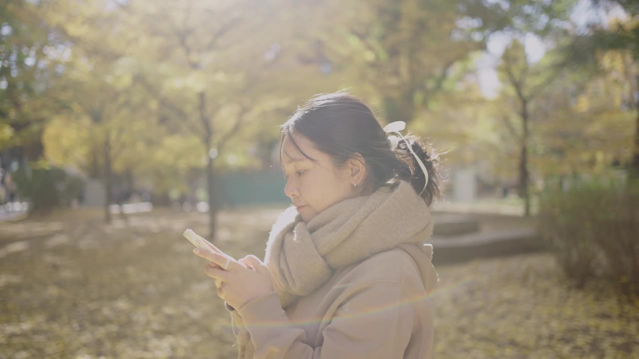 秋天，一名年轻女子在城市公园拿着智能手机拍摄照片或视频，将令人惊叹的景色分享到社交媒体上。视频下载