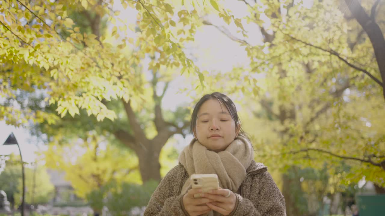 秋天，一名年轻女子在城市公园拿着智能手机拍摄照片或视频，将令人惊叹的景色分享到社交媒体上。视频下载