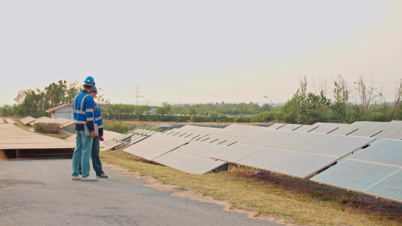 两名身穿黄色背心的男子走在一排太阳能电池板旁边的小路上。进步和创新的概念，因为工人可能会参与太阳能电池板的安装或维护视频下载