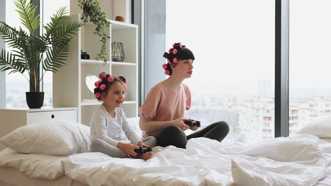一头卷发的母女坐在家里的床上玩电子游戏。视频下载